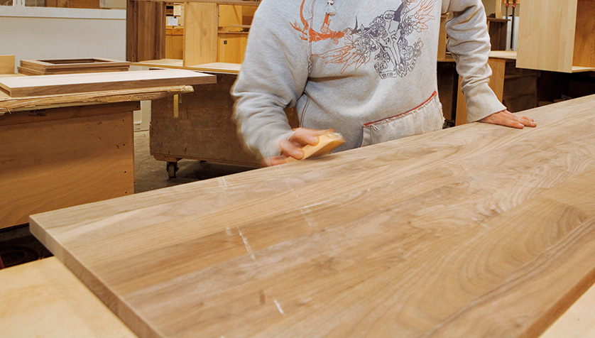 An artisan's hands are pictured sanding a board in a workshop.